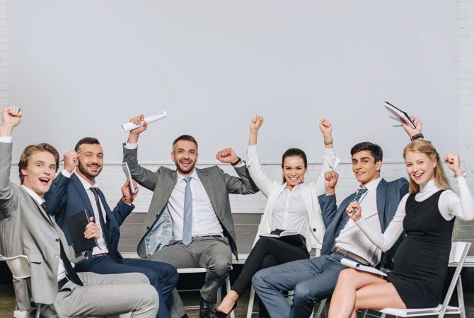 happy-businesspeople-with-raised-hands-sitting-on-chairs-at-training-in-hub.jpg